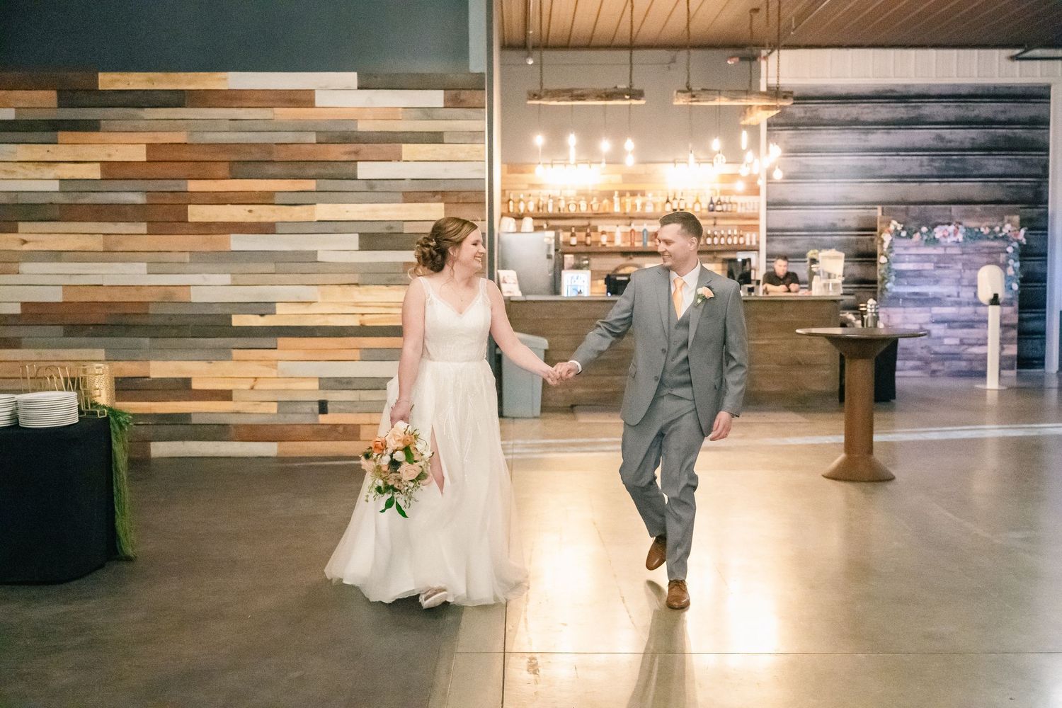 Bride and groom walk hand in hand past reclaimed wood wall at reception.