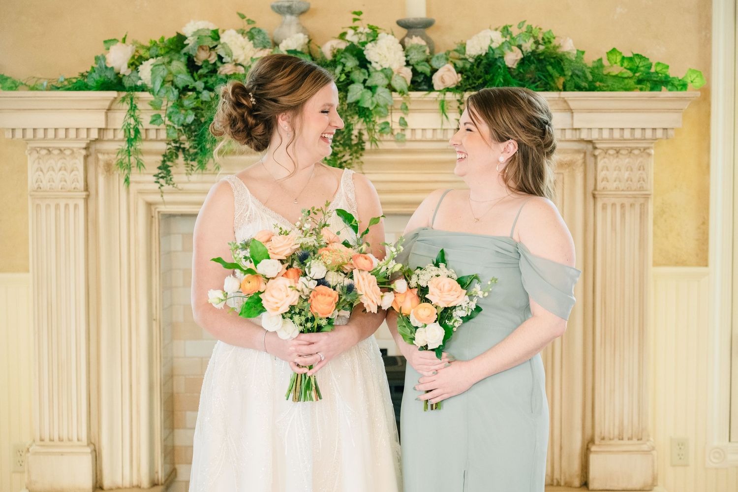 Bride and bridesmaid in sage off-shoulder dress holding peach and white bouquets in front of a floral fireplace.