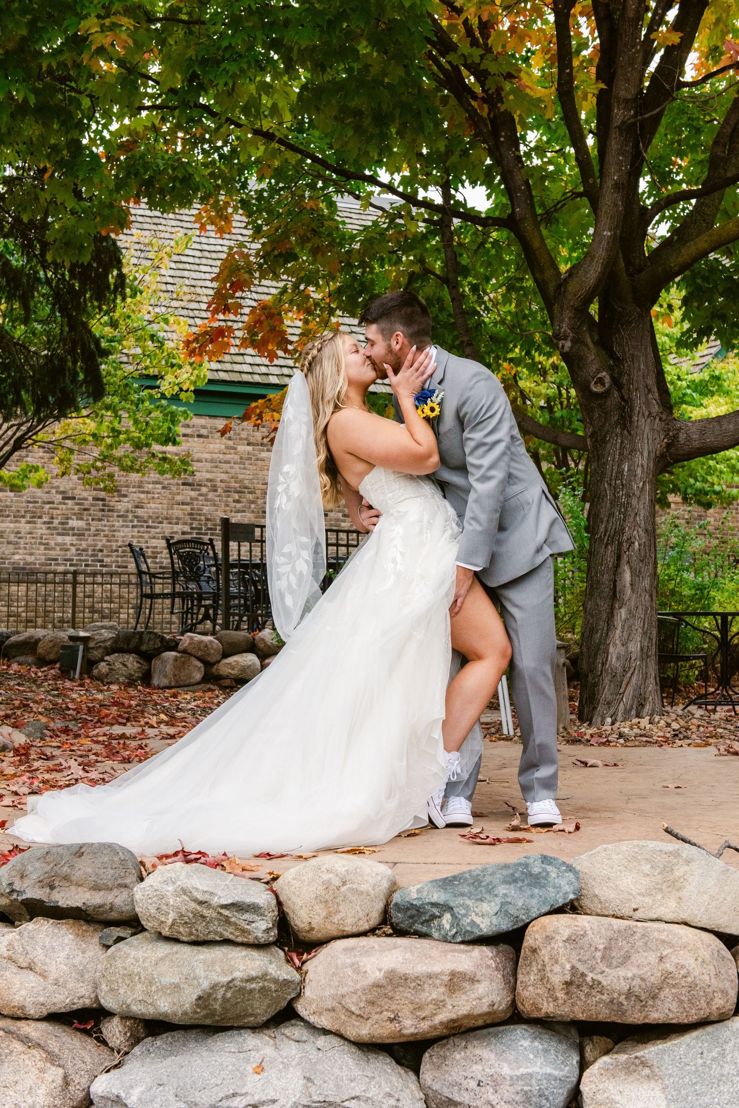 Central MN Wedding Photos | Bride and groom sharing a romantic kiss on a rock wall surrounded by colorful autumn foliage.