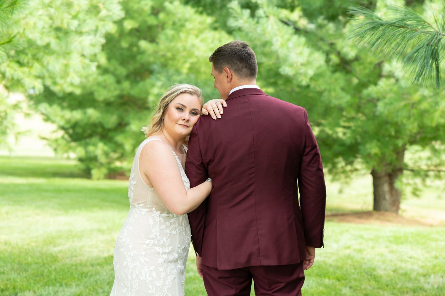 Central MN Wedding Photos - Bride in white gown rests hand on groom's burgundy suit shoulder outdoors with lush green trees in background.