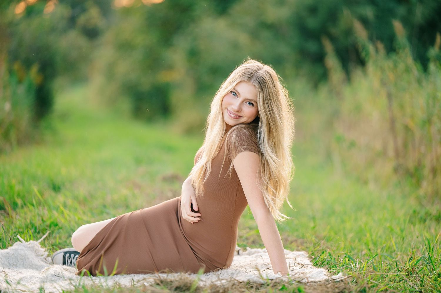 Central MN Senior Photography - Young blonde woman in brown dress sitting gracefully on grass in a sunlit outdoor field, smiling warmly.