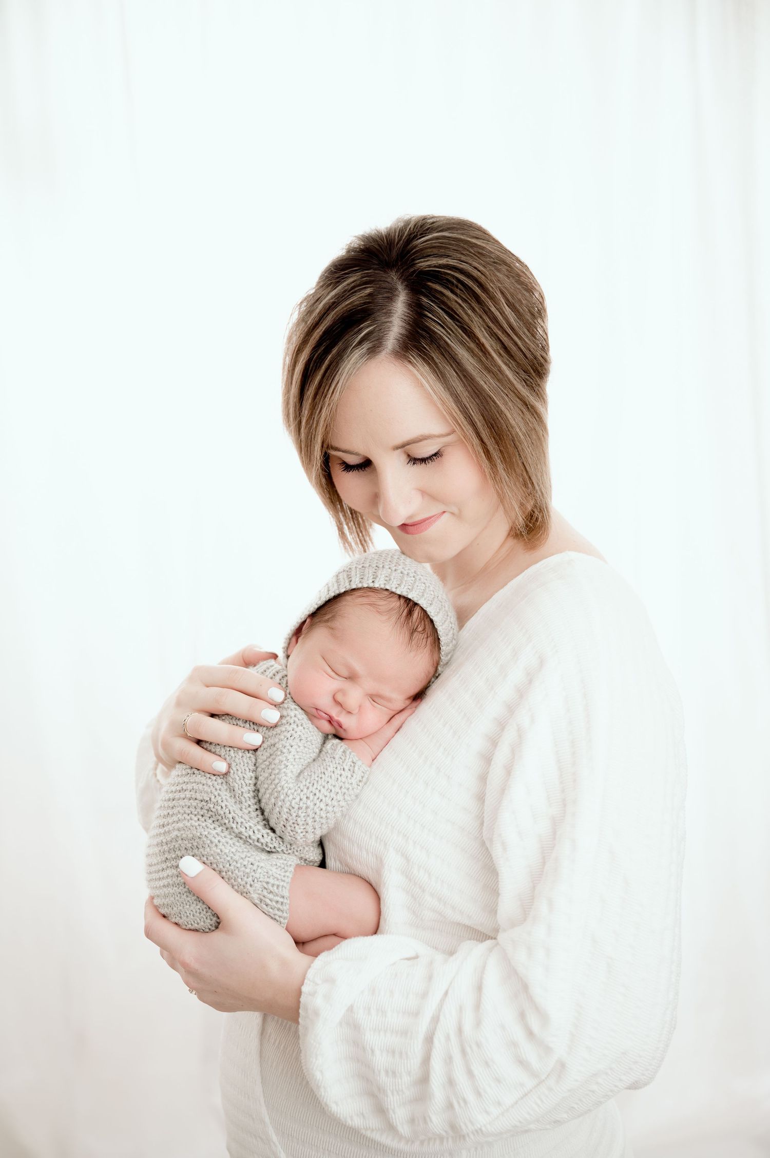 St Cloud newborn Photos of A smiling mother in a white sweater tenderly holds her sleeping newborn baby wrapped in a gray knit outfit.