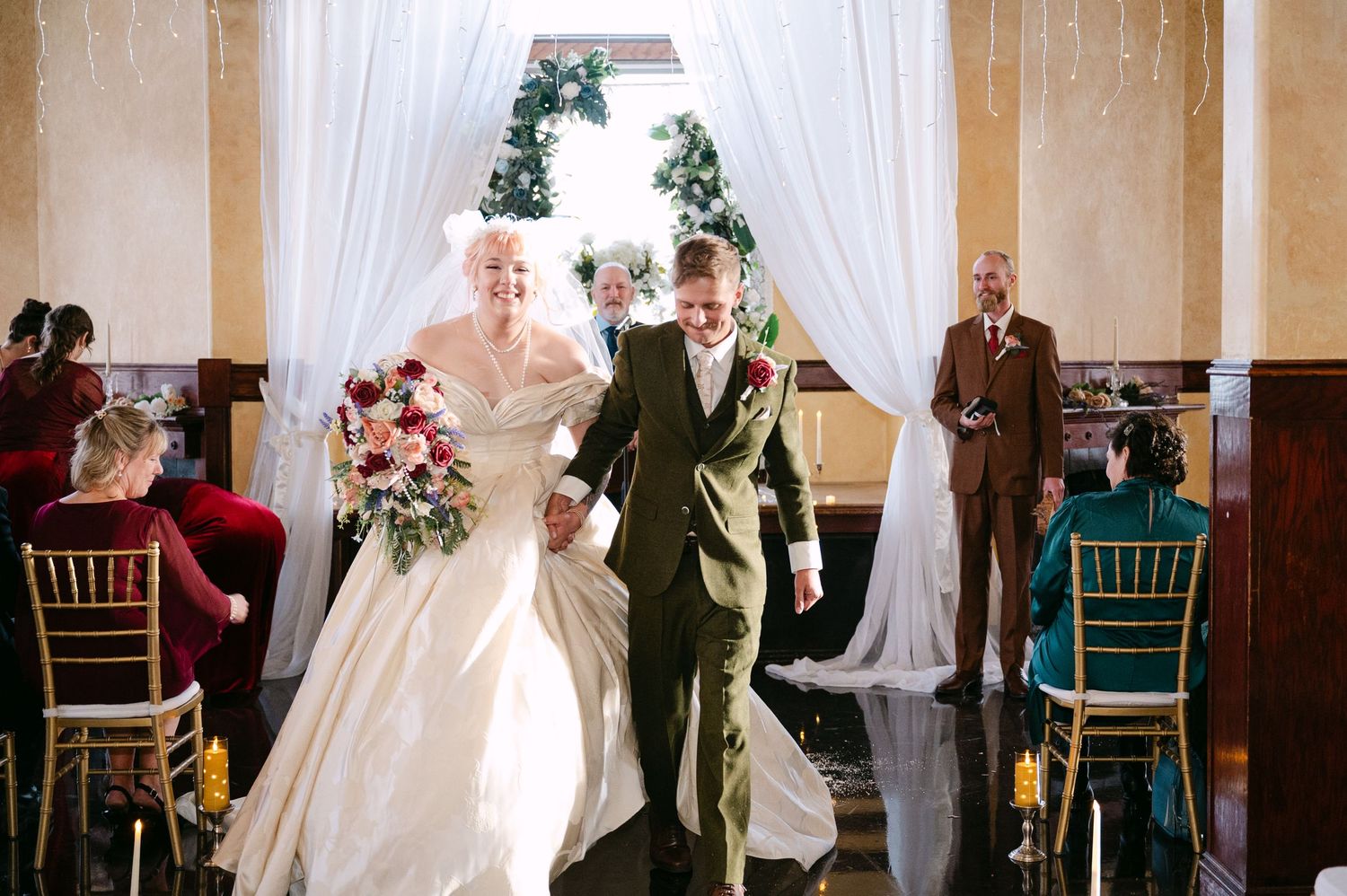 Central MN Wedding Photos | Newlyweds walk joyfully down aisle after ceremony, bride holding red bouquet, groom in olive suit.