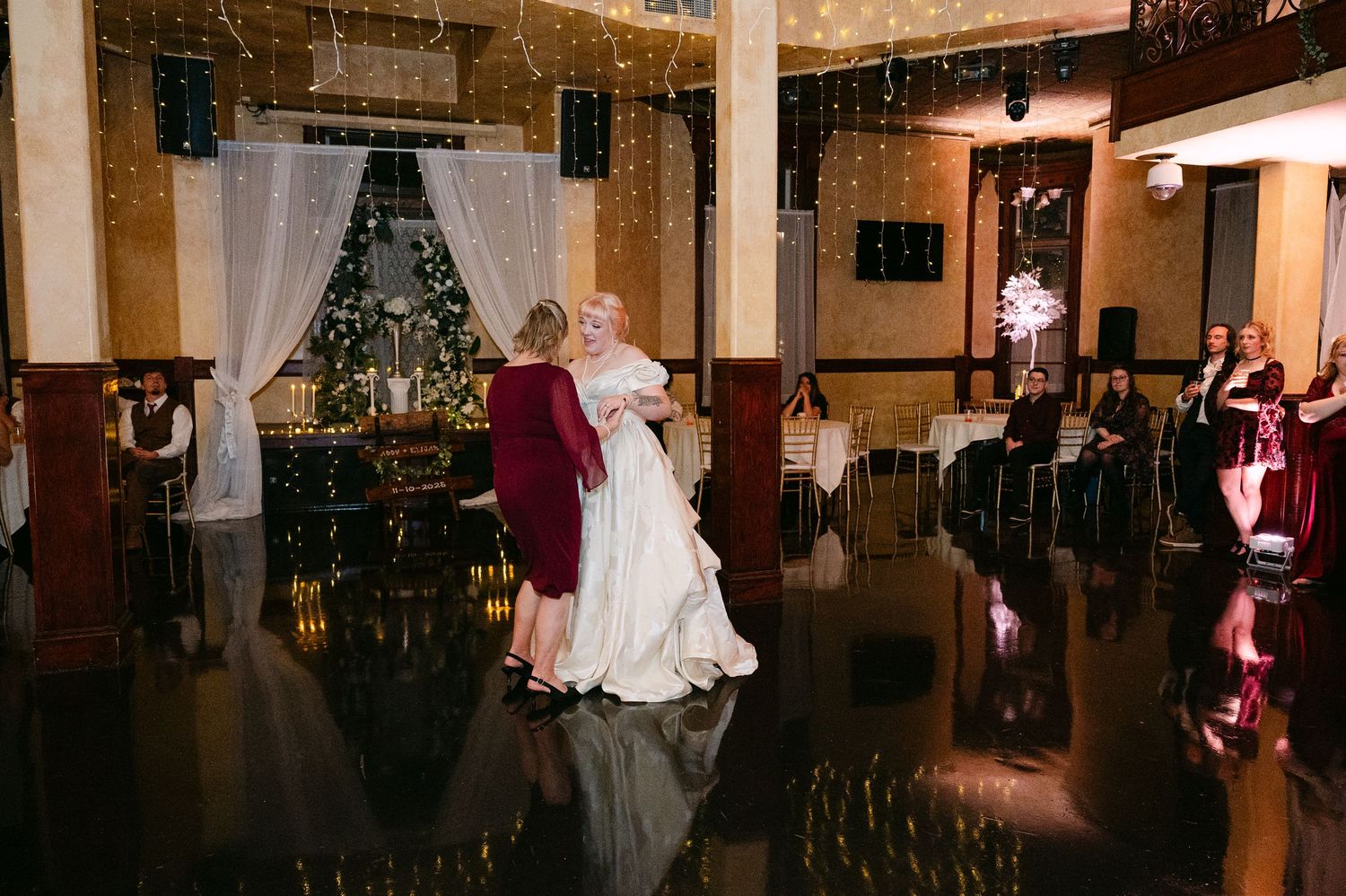 Central MN Wedding Photos | Bride in ivory gown shares emotional dance with guest in burgundy dress on black reflective dance floor.