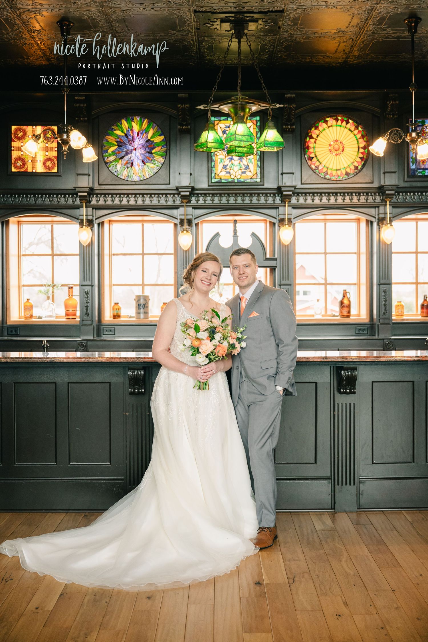 Wedding couple stands in front of vintage stained glass windows in historic venue with warm lighting and wood floors.