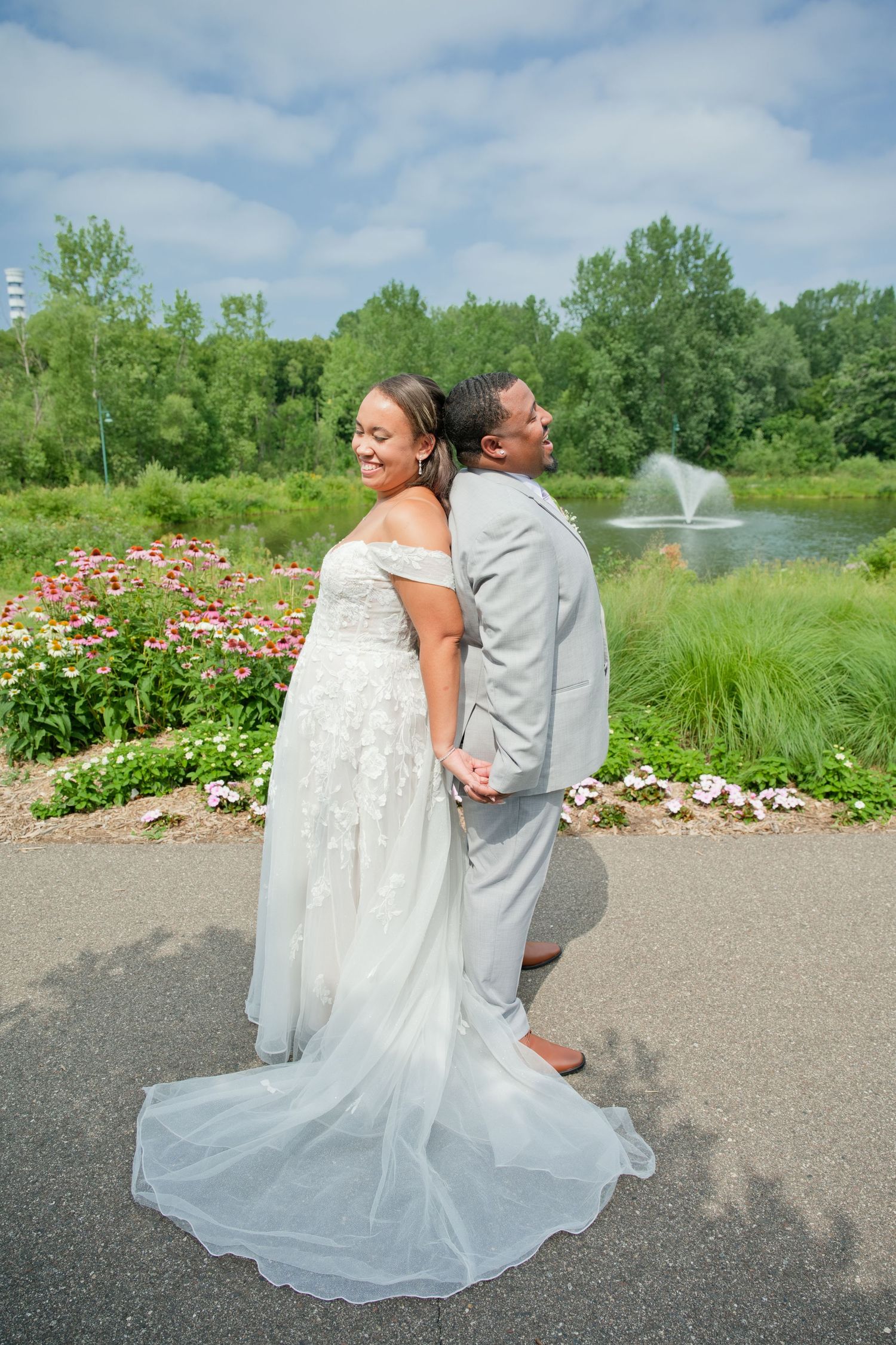 Bride and groom stand back-to-back holding hands near garden fountain on wedding day.