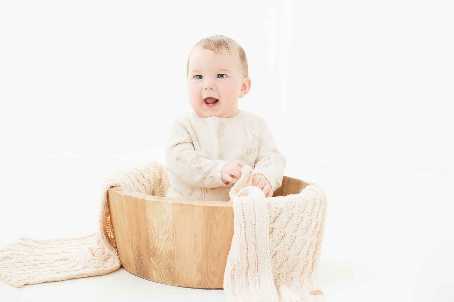 Baby in cream outfit sitting in wooden bowl with knit blanket on white background.. Minnesota milestone photographer