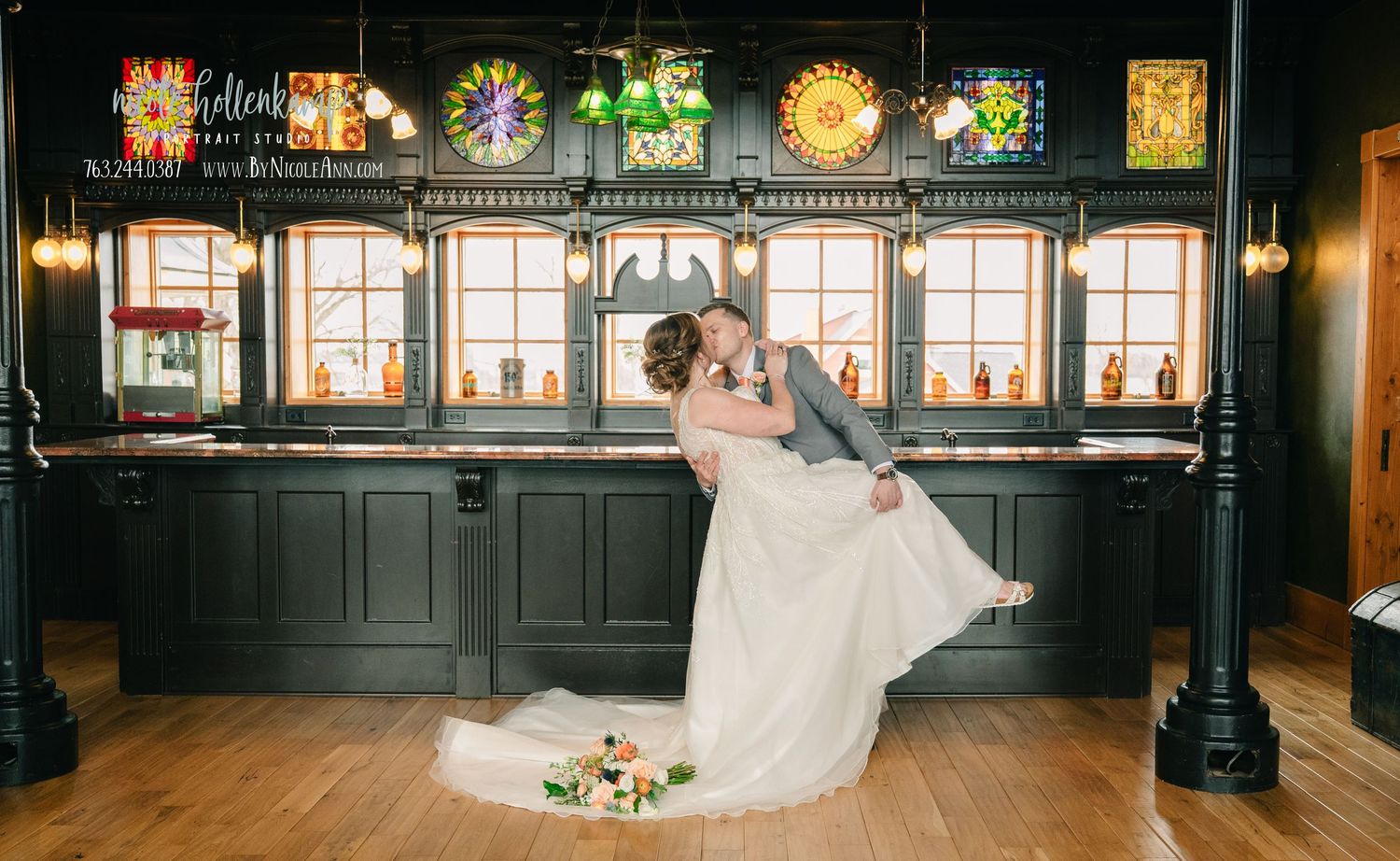 Bride and groom share a romantic dip kiss in front of an elegant dark wood bar with stained glass decor. Crooked Willow Osakis