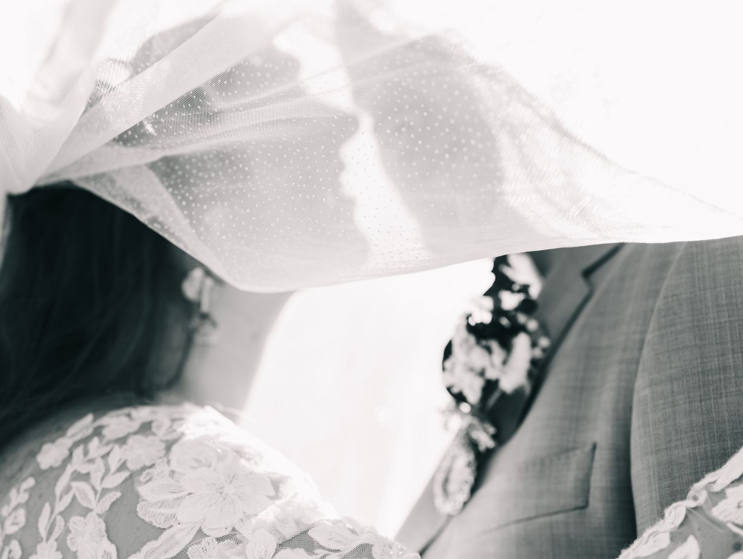 Black and white romantic photo of bride and groom sharing a kiss beneath a flowing wedding veil.