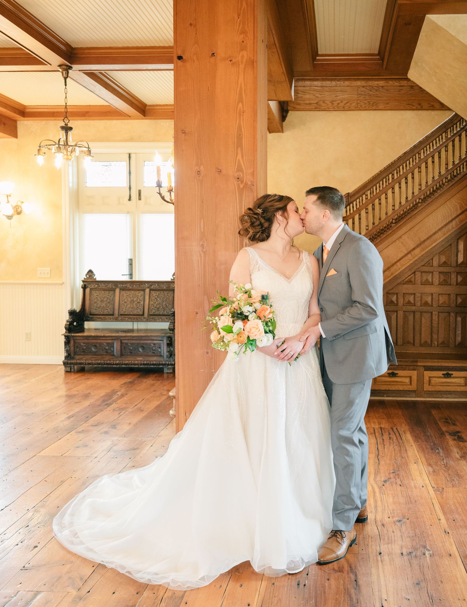 Bride and groom share a kiss in an elegant wooden venue, bride holding a colorful bouquet beside a grand staircase.