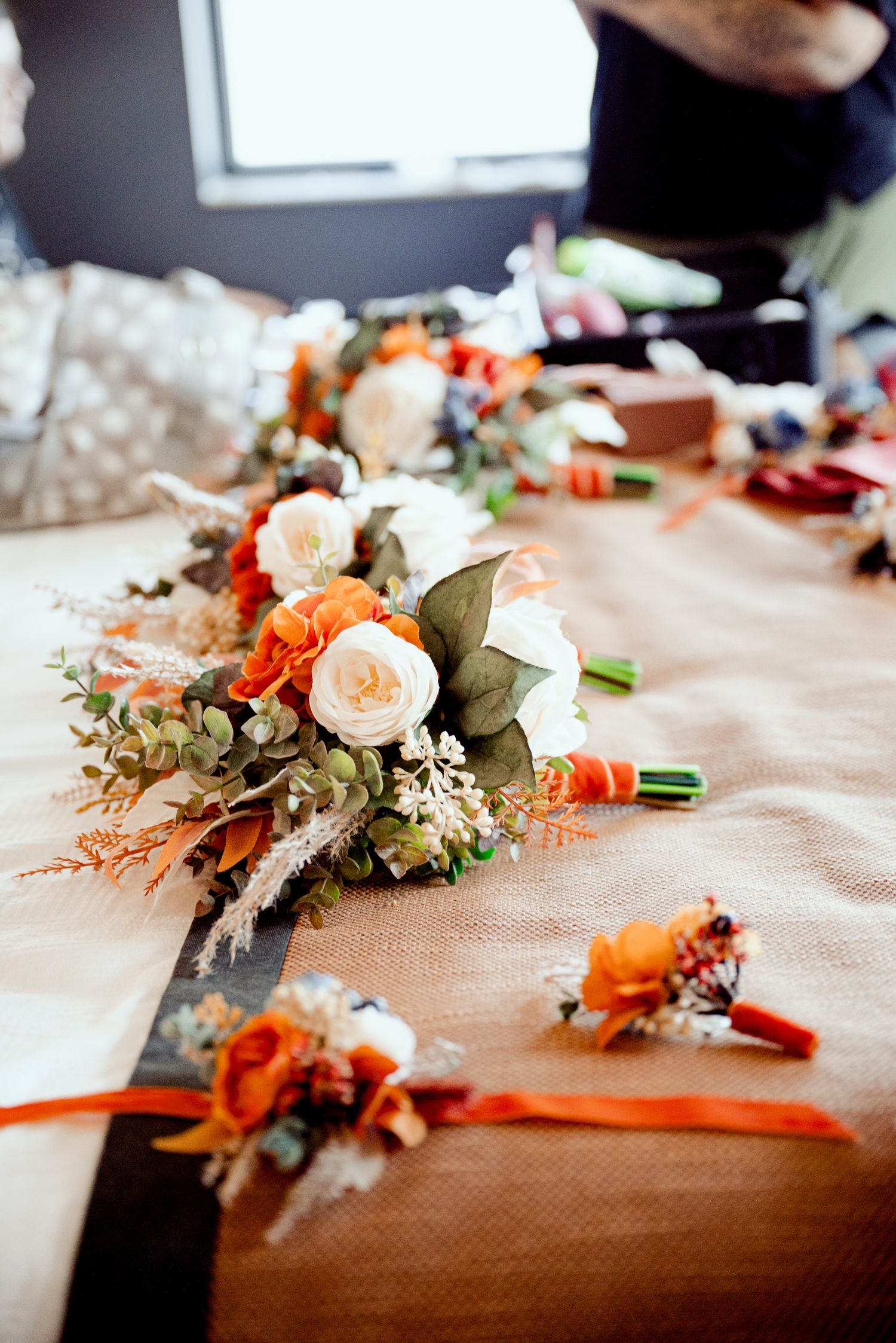 Multiple floral wedding bouquets with orange and white flowers laid out on a beige tablecloth during wedding preparations.