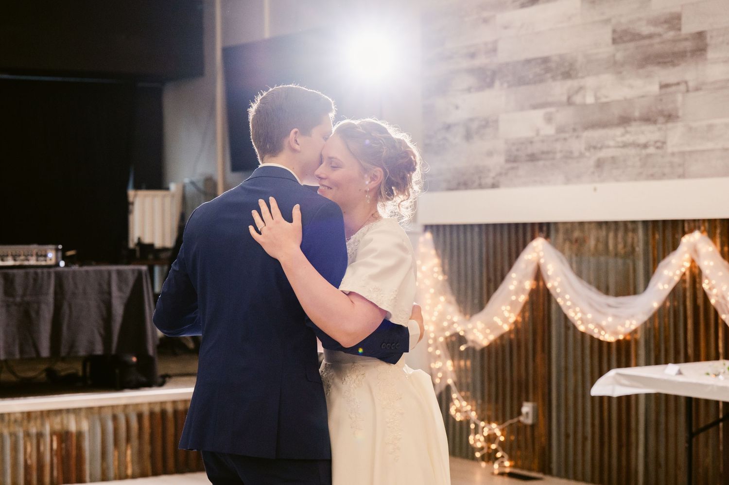 A couple embraces during their first dance with string lights and rustic decor in the background.