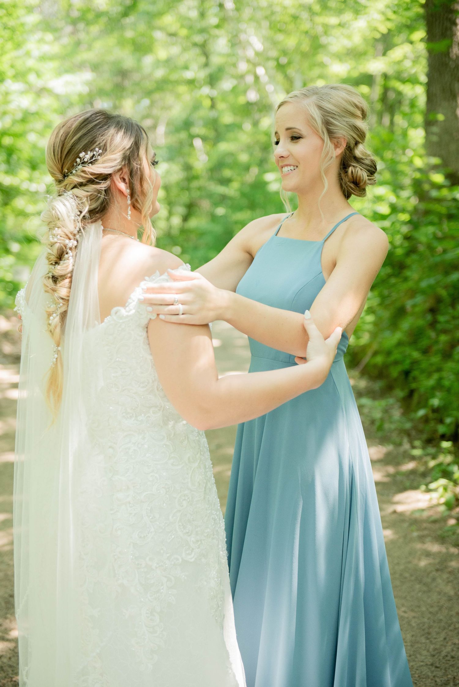 A bridesmaid in a blue dress shares a tender moment with the bride wearing a flowing white gown in a garden setting. Zimmerman MN Weddings