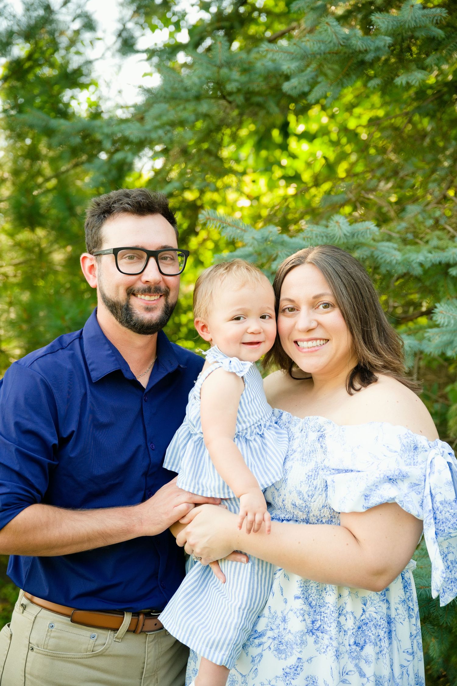 A happy family of three poses outdoors, parents in blue outfits holding their smiling toddler among green trees.