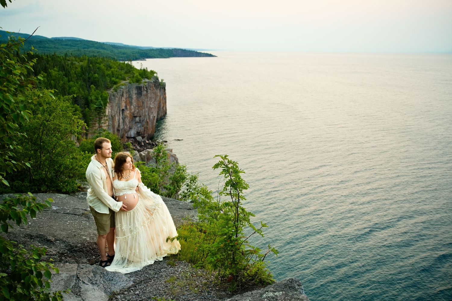Bride and groom stand on cliff edge overlooking vast blue lake surrounded by lush greenery.