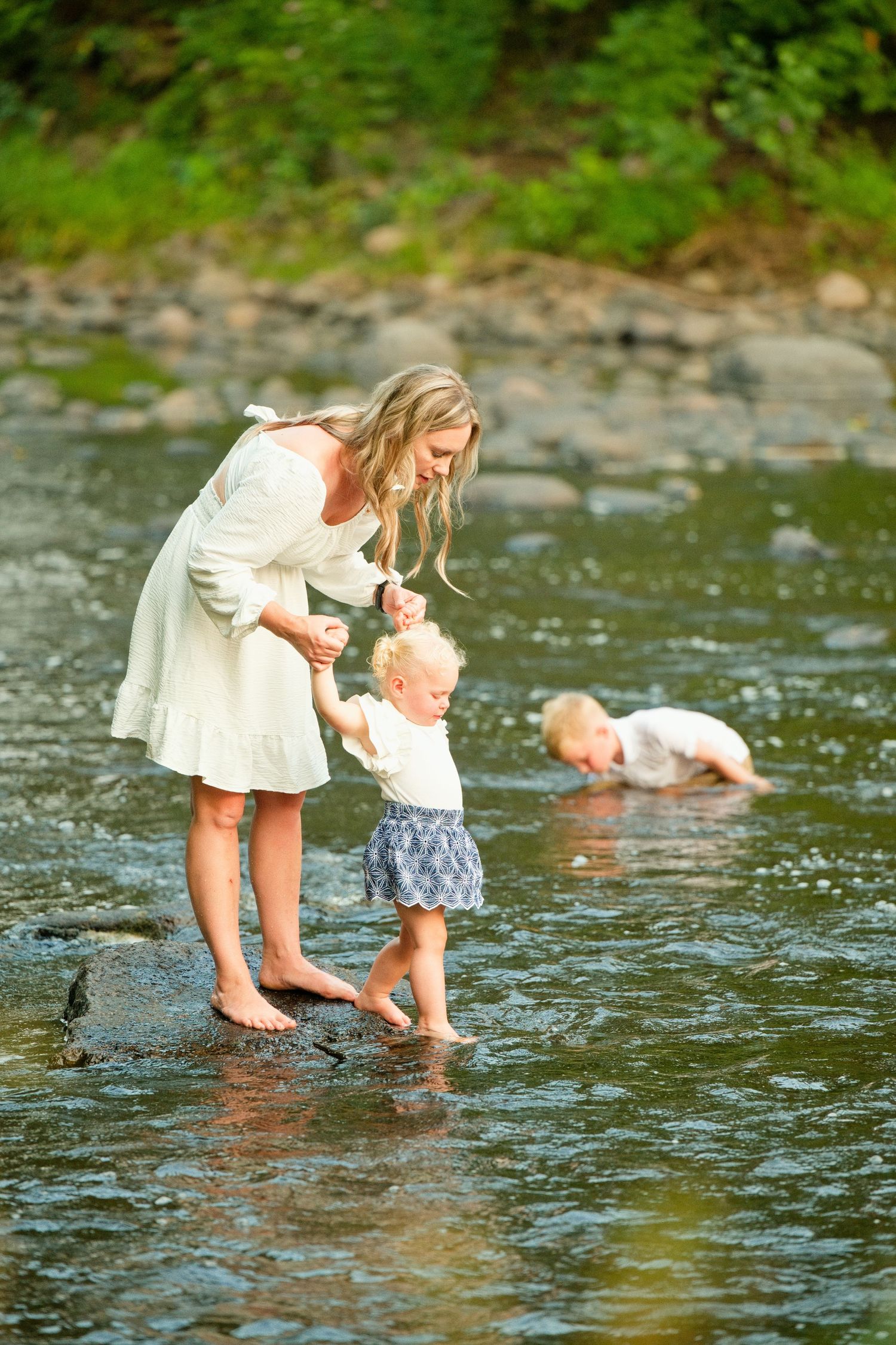 A mother in a white dress helps her toddler walk in a shallow rocky stream while another child plays nearby.