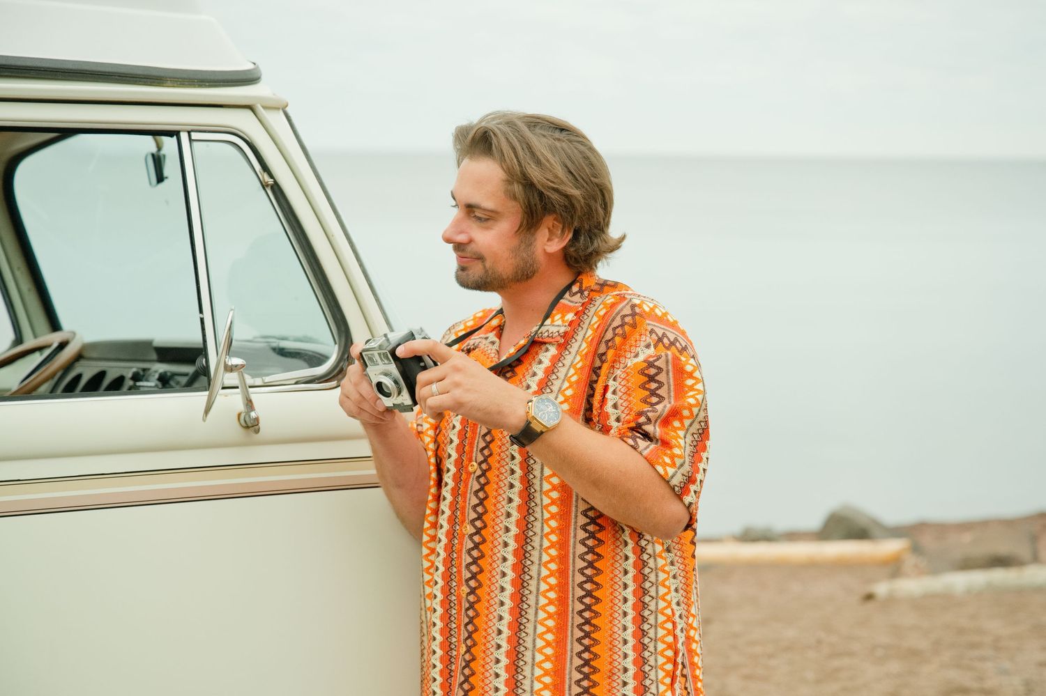 Man in orange patterned shirt holding vintage camera beside retro VW van on beach.