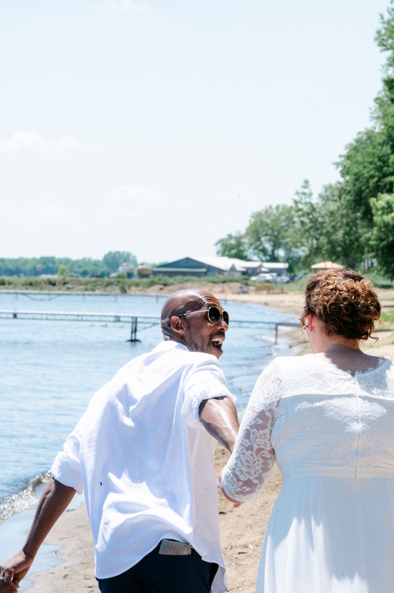 Happy interracial couple in wedding attire walking along sandy beach shoreline on a bright sunny day.