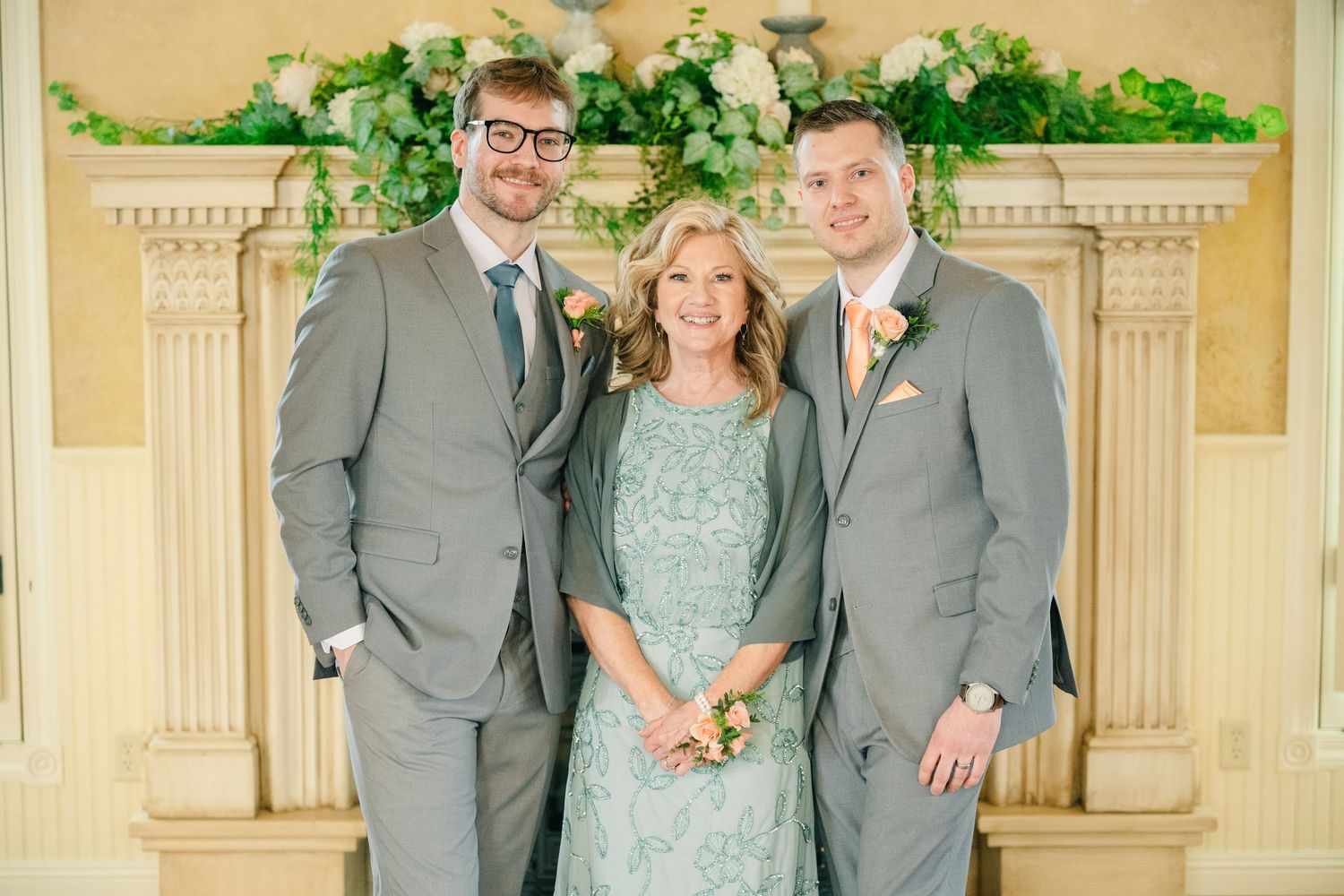Two groomsmen in gray suits flanking a woman in a sage green lace dress in front of floral fireplace.