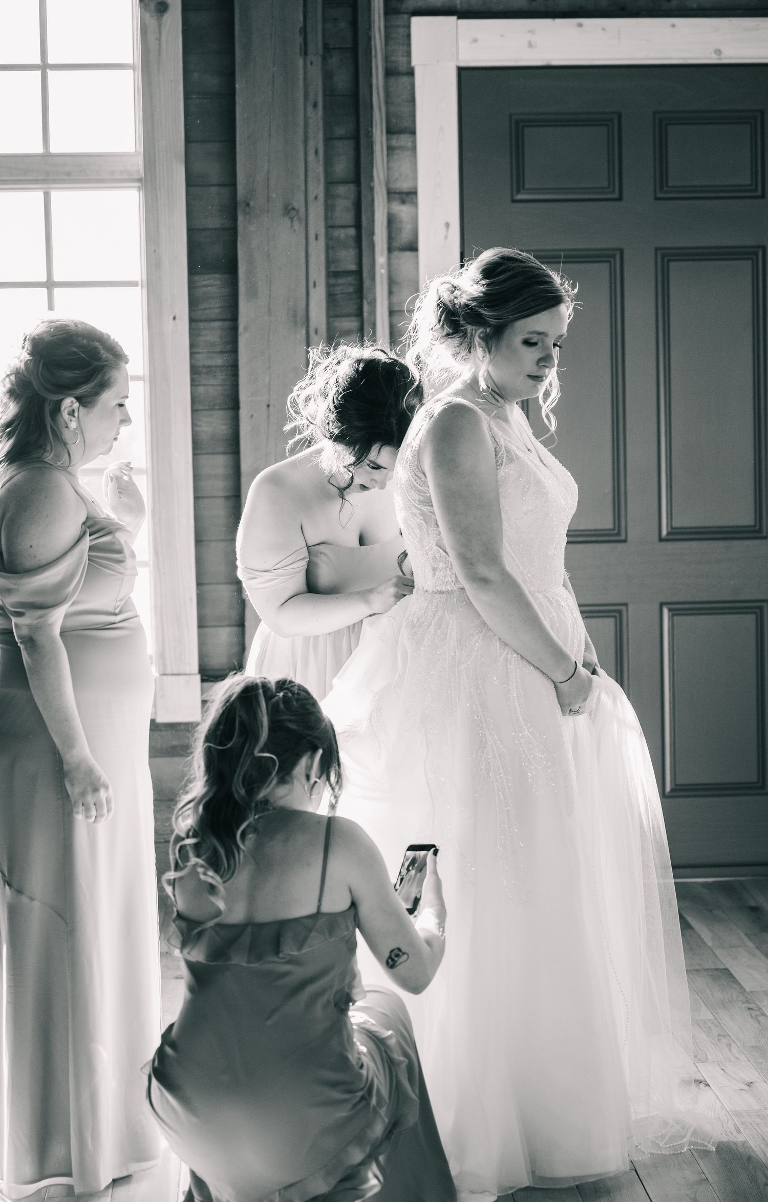 Bridesmaids help bride with her wedding gown in a black and white getting-ready photo.