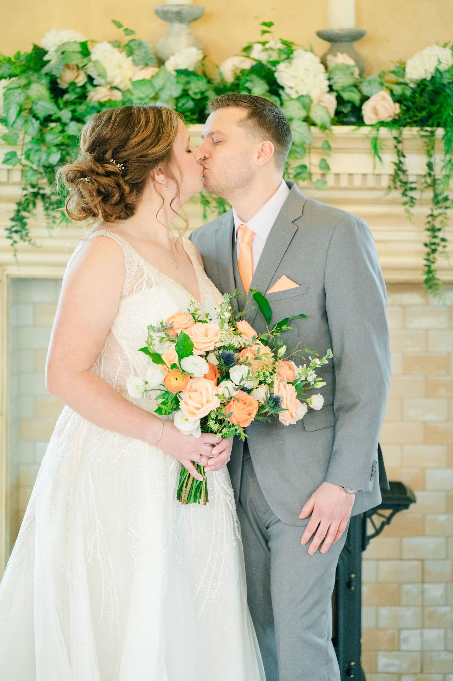 Bride and groom share a kiss holding a peach rose bouquet in front of a floral fireplace mantle.