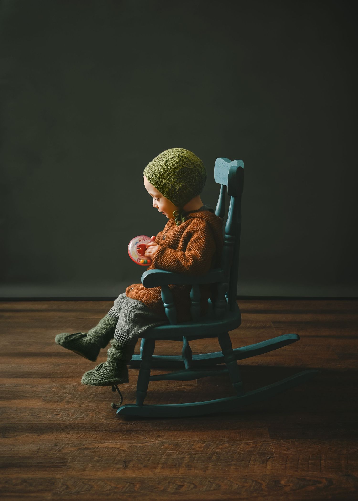 A small figure in a knit cap and brown coat sits in a blue painted wooden rocking chair on a hardwood floor.