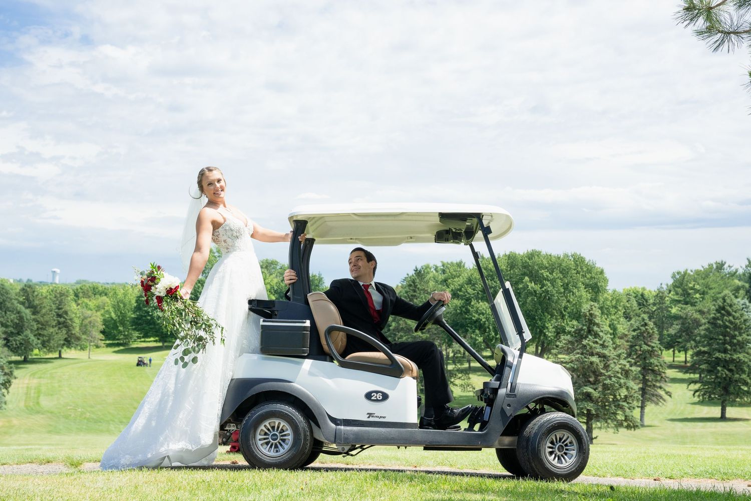 Smiling bride in white gown poses beside groom driving a golf cart on a lush green golf course.
