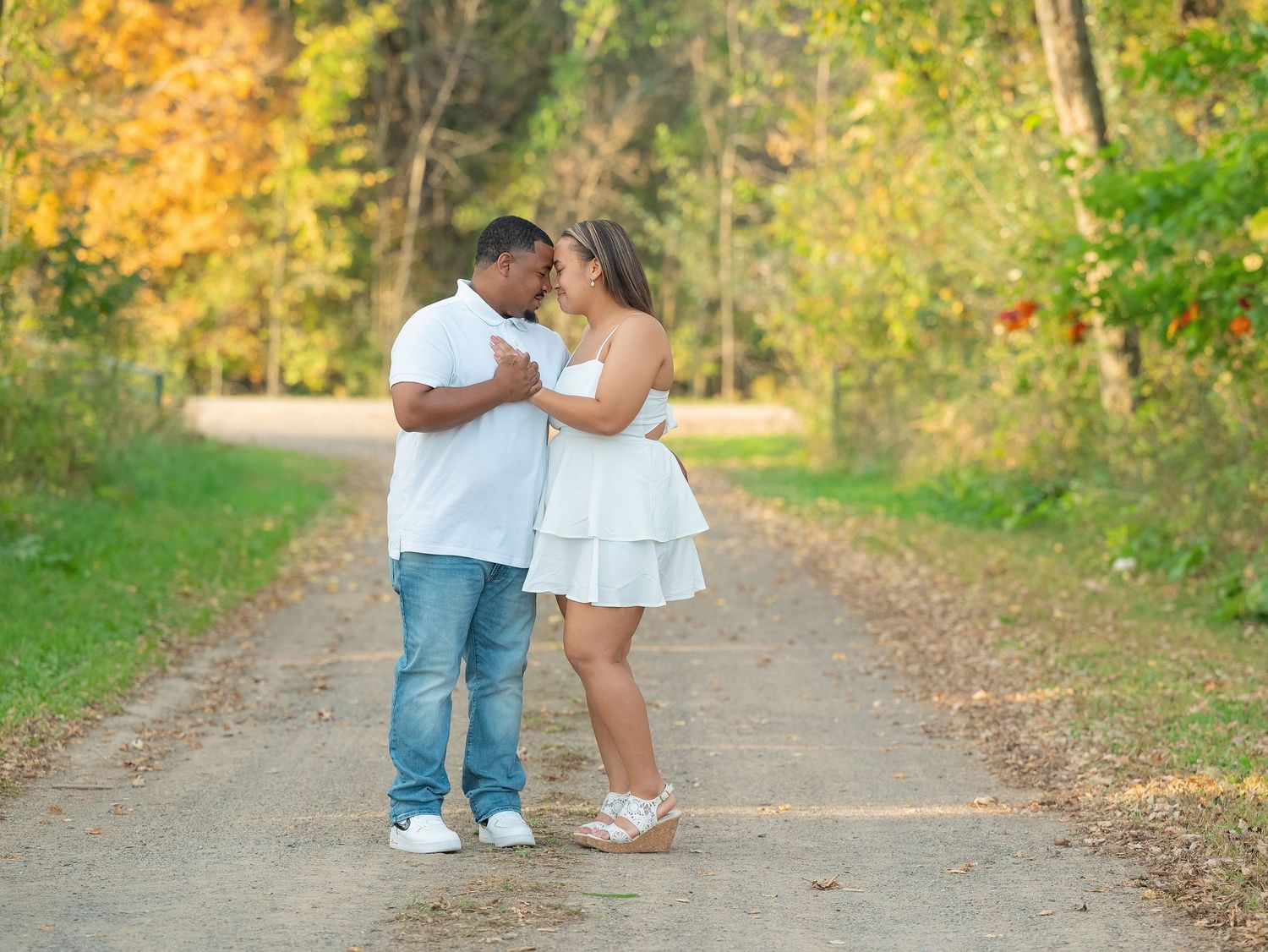 A couple stands together on a sunlit autumn path surrounded by golden foliage and trees.