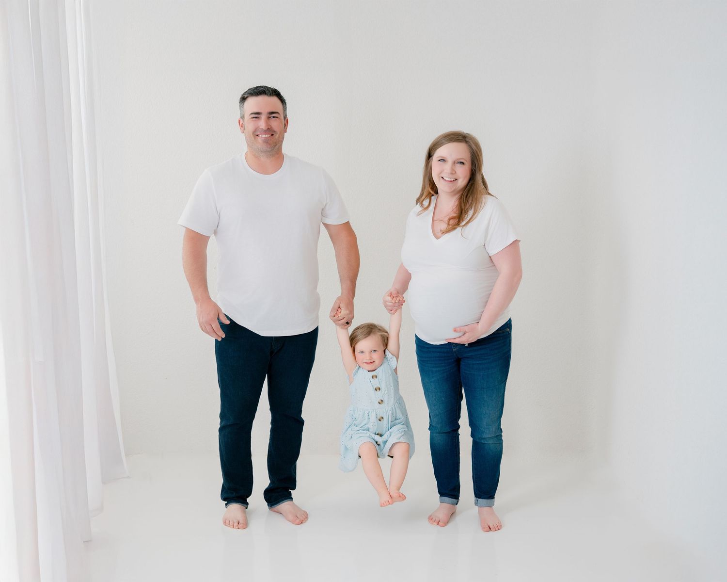A family of three wearing casual white tops and jeans poses together against a white backdrop in a bright studio setting.