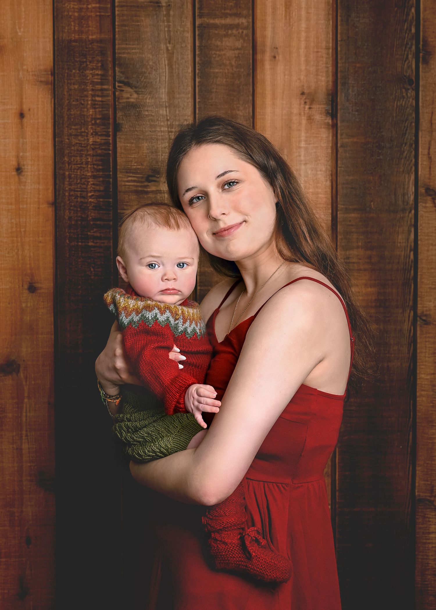 A parent in a red dress holds a baby wearing a festive green and red knit sweater against a wooden wall backdrop.