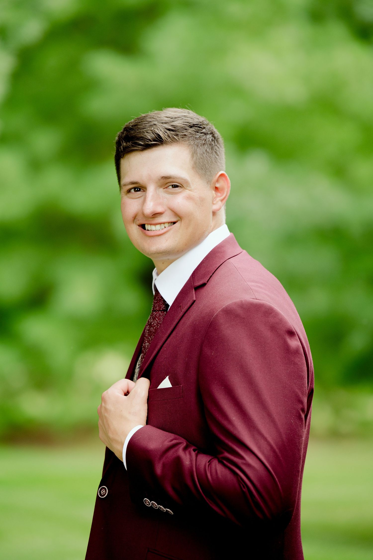 A groom poses outdoors in a burgundy suit jacket against a blurred green background.