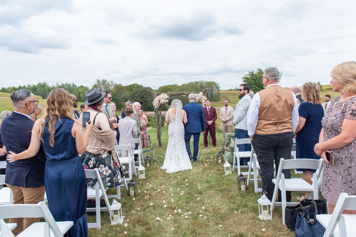Wedding guests seated in white chairs for an outdoor ceremony in a rustic countryside setting.