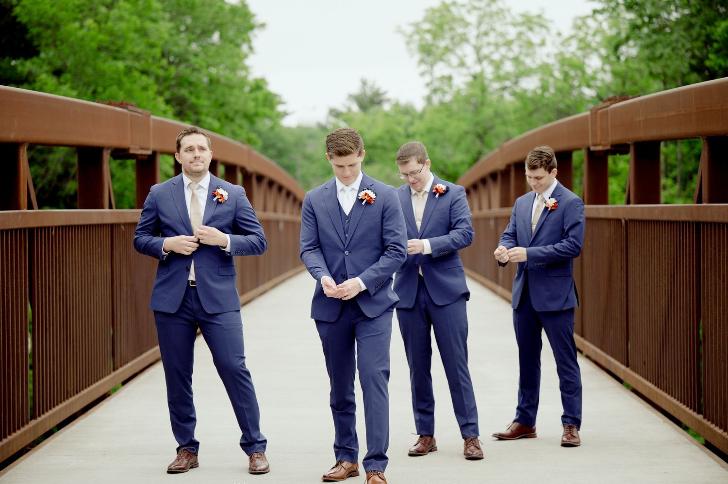 Four groomsmen in matching navy suits pose casually on a wooden bridge surrounded by greenery.