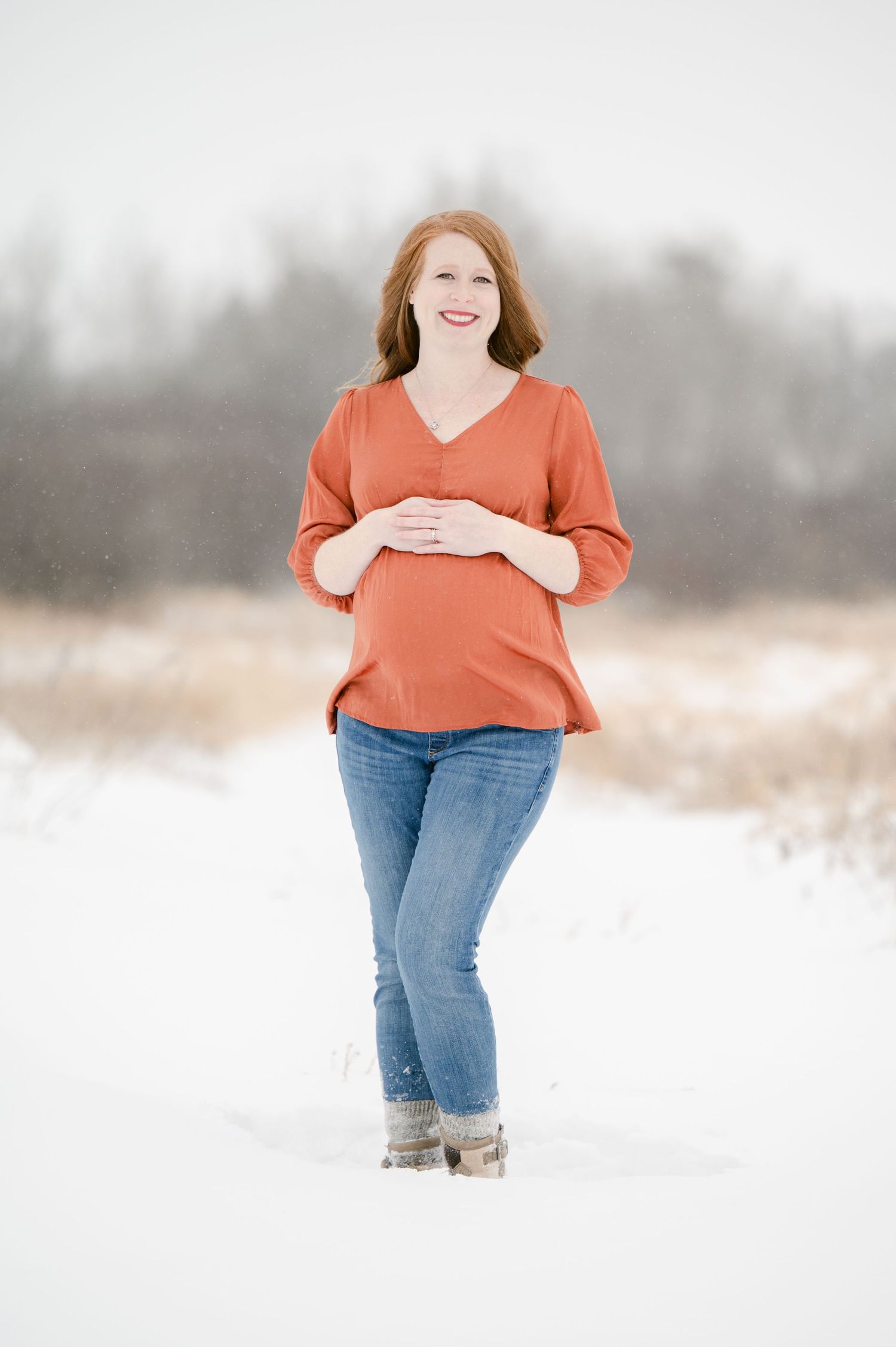 A person in an orange sweater and jeans poses confidently in a snowy winter field.