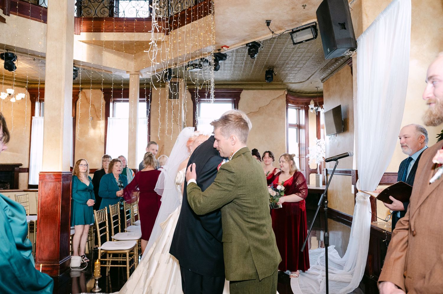 Central MN Wedding Photos | Bride and groom share first kiss at wedding altar surrounded by guests in decorated venue.