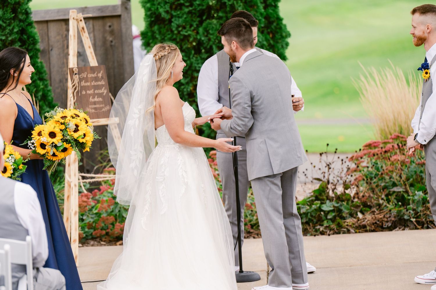 Central MN Wedding Photos -Bride and groom exchanging vows at an outdoor wedding ceremony with sunflower bouquets and guests seated in rows.