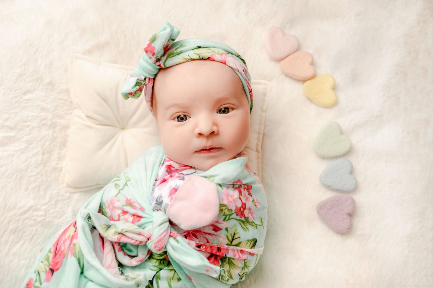 St Cloud newborn Photos of Newborn baby girl swaddled in floral wrap with bow headband, lying next to pastel heart decorations on blanket.