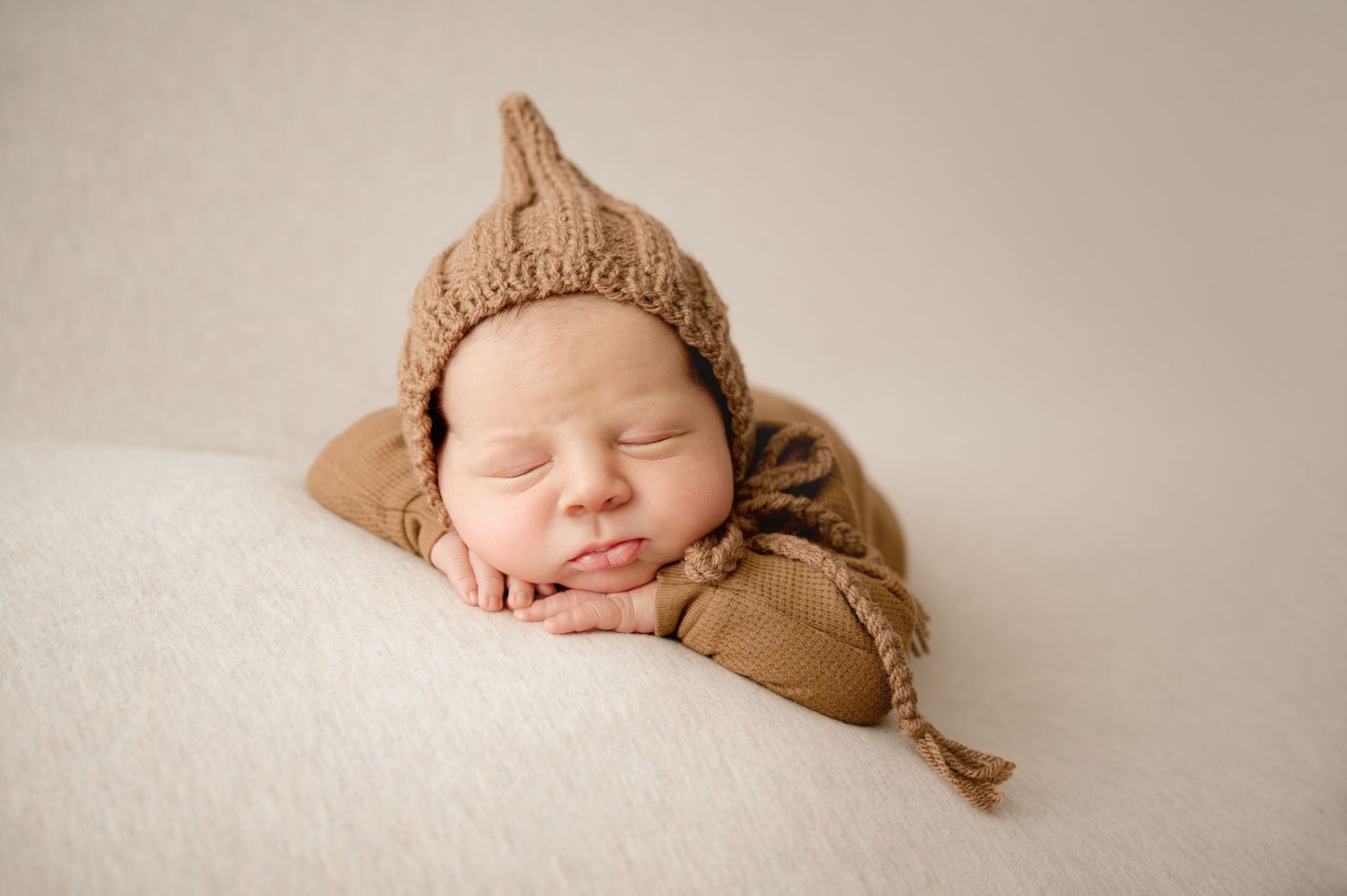 Sleeping newborn baby wearing a cozy brown knit bonnet and romper, posed on a soft white blanket.