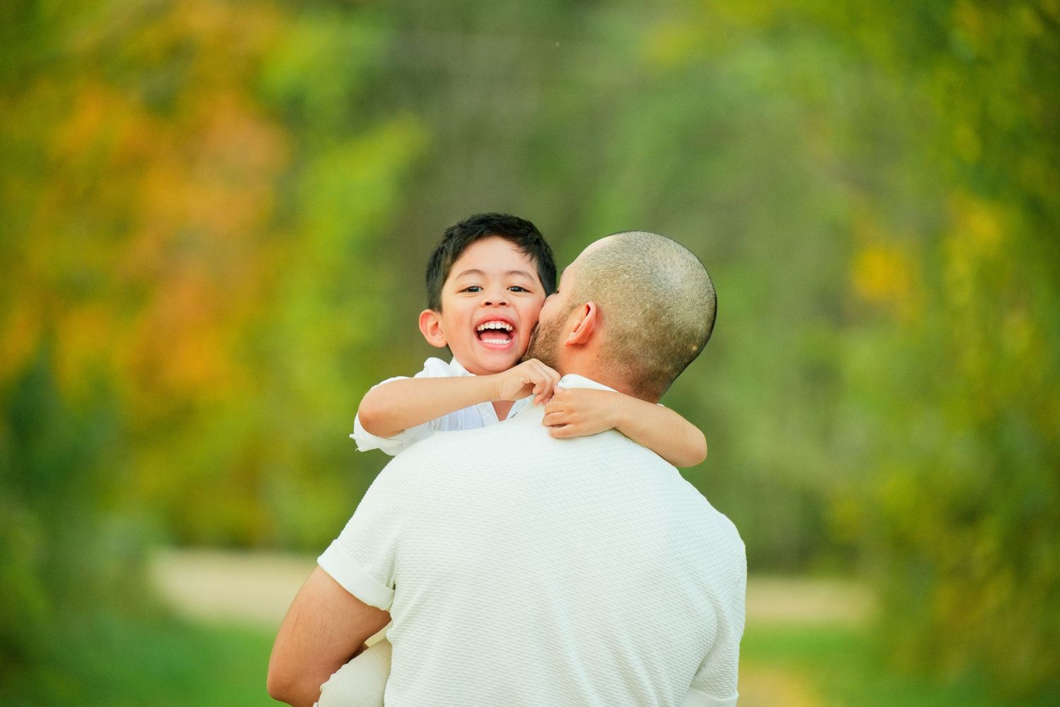 A joyful young boy laughs while being held by a man outdoors in a lush green park setting.