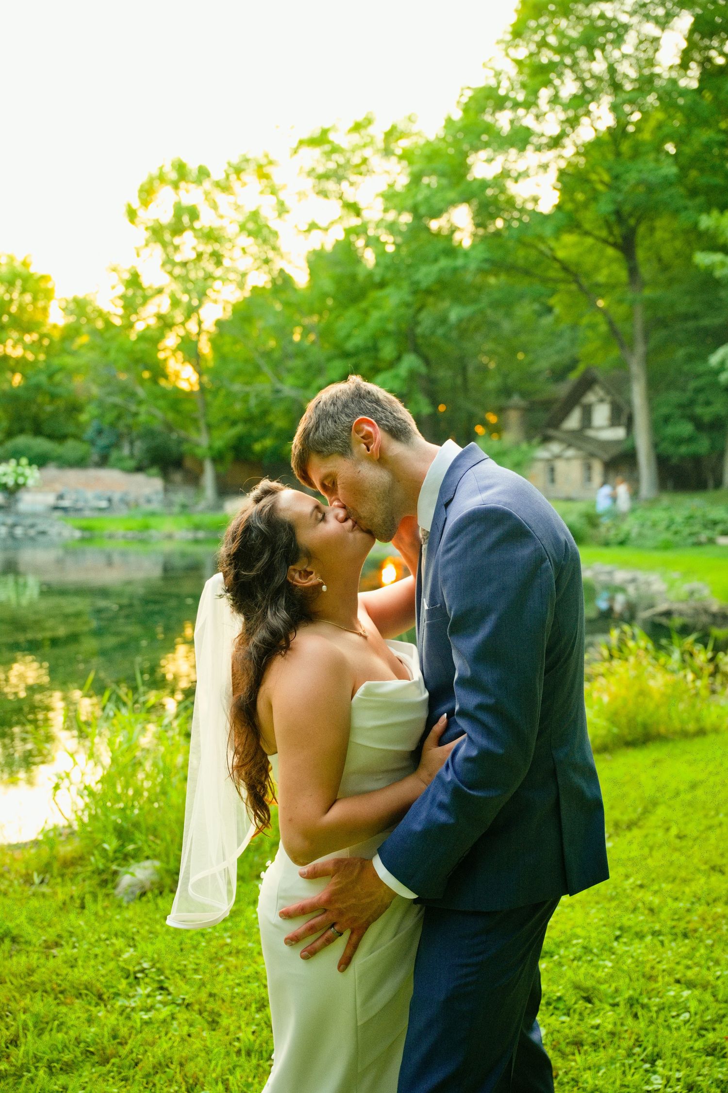 Bride and groom sharing a romantic kiss outdoors near a pond, surrounded by lush green trees at golden hour.