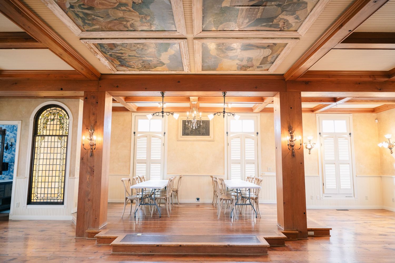 Elegant banquet hall with painted ceiling, wood beams, stained glass window, and tables set for an event.