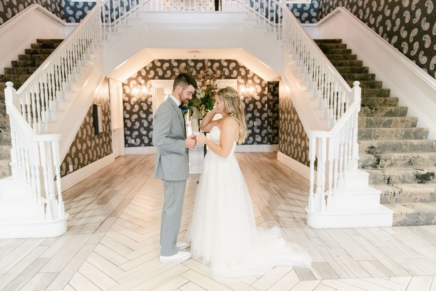 Central MN Wedding Photos | Bride and groom share first look moment in elegant venue foyer with grand white staircases.