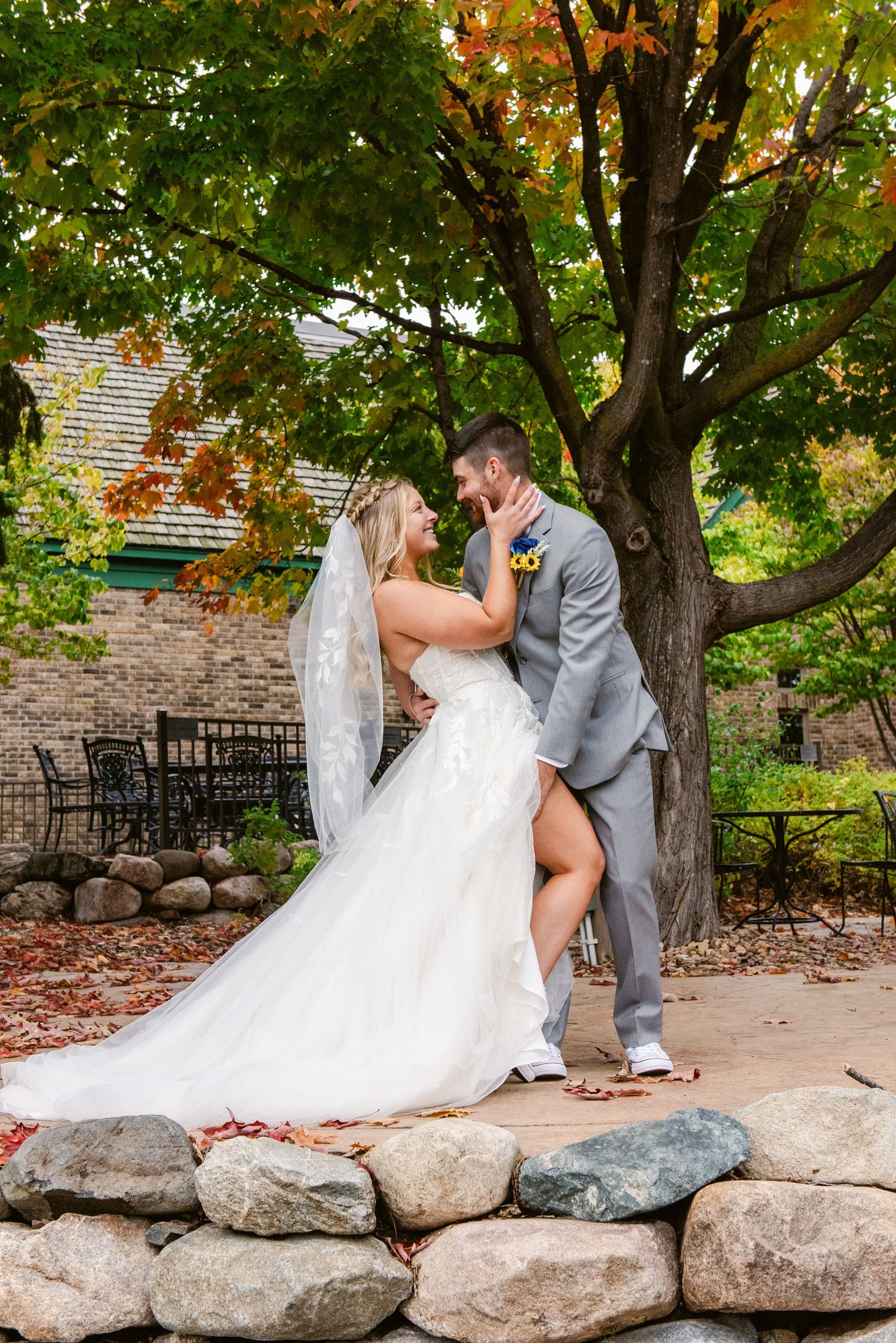 Central MN Wedding Photos | Bride and groom share romantic moment under autumn tree on stone wall outdoors.