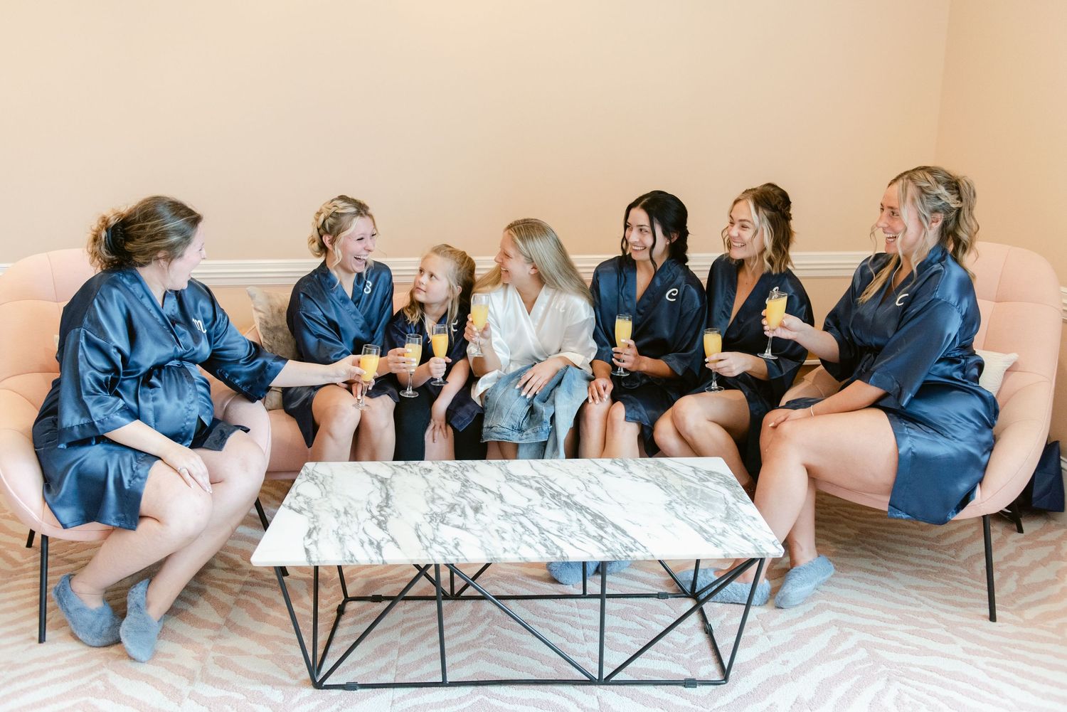 Central MN Wedding Photos | Bridesmaids in satin robes toasting with champagne on a couch, celebrating before a wedding ceremony.