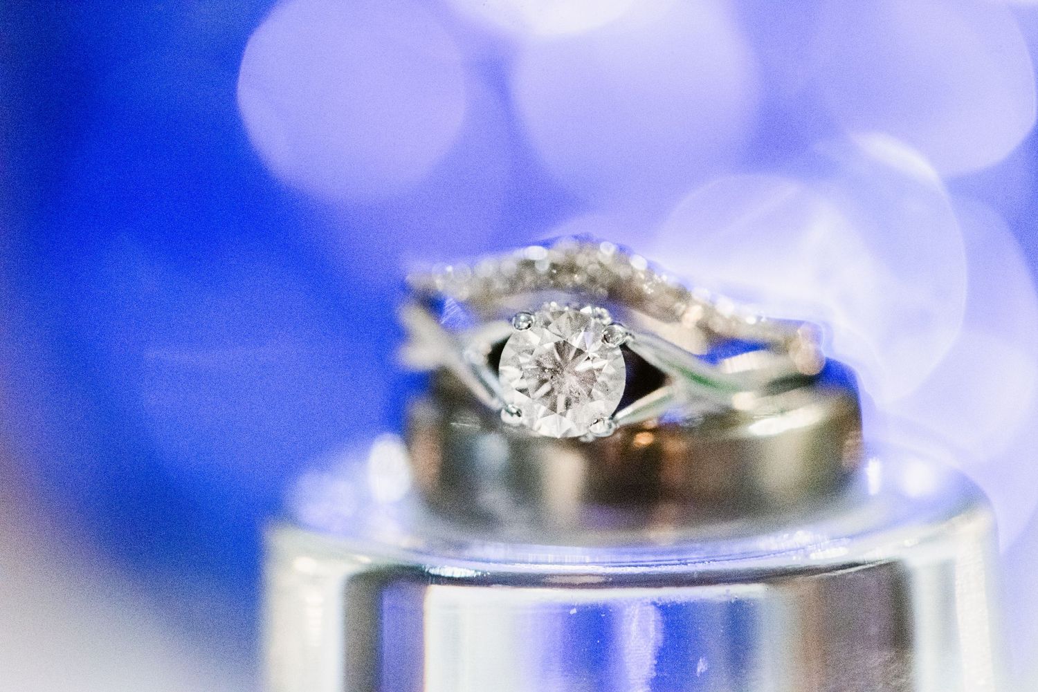 Central MN Wedding Photos - Close-up of diamond engagement ring and wedding bands stacked on reflective surface with blue bokeh.
