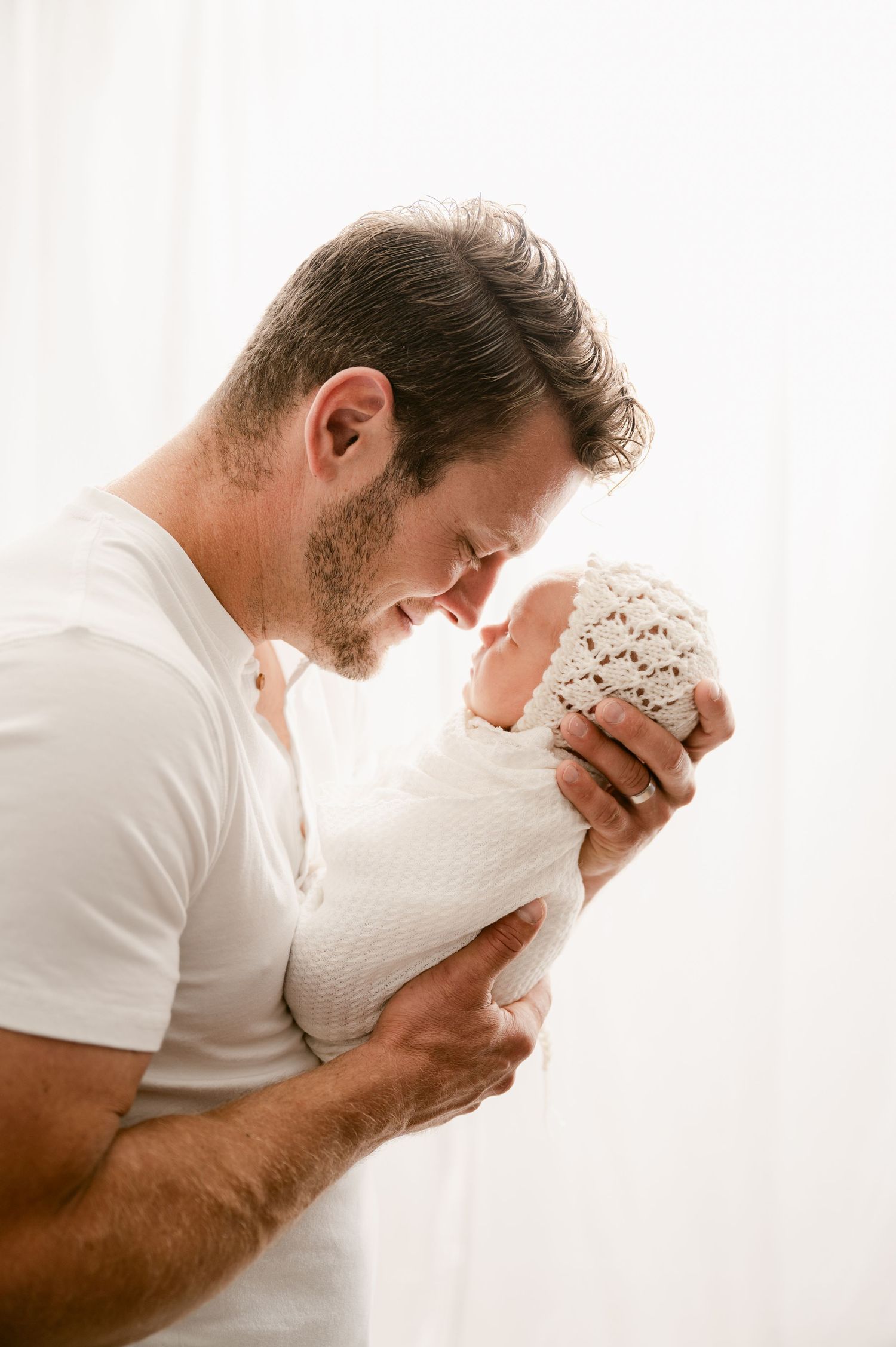 A father tenderly holds his swaddled newborn baby close, touching foreheads in a bright, airy white studio setting.