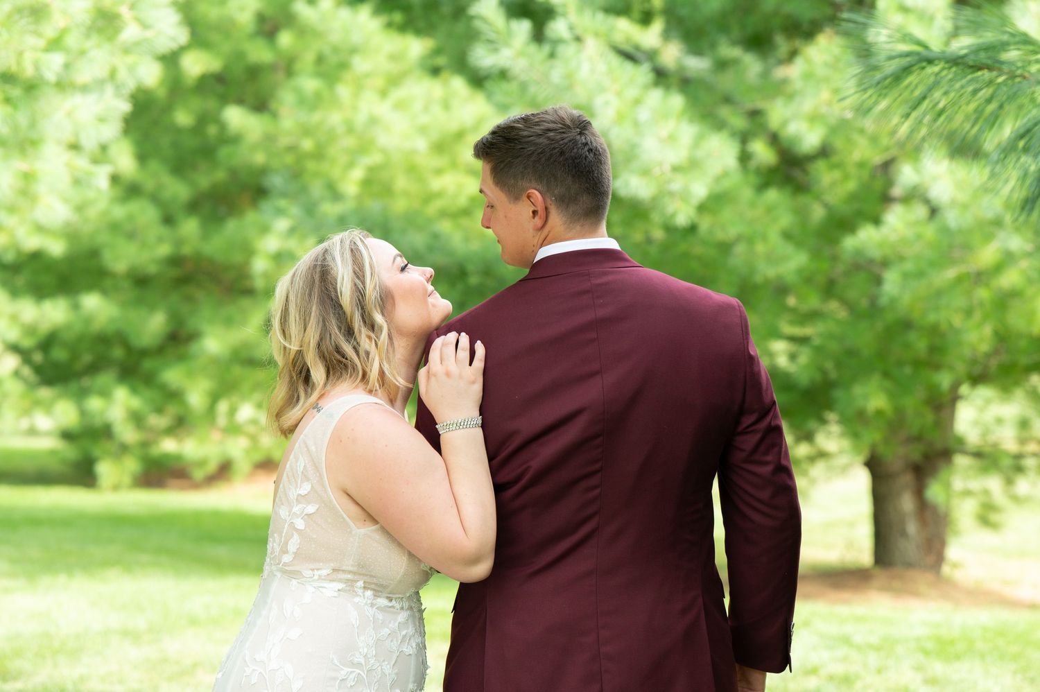 Central MN Wedding Couple sharing romantic moment outdoors, bride in lace gown touching groom's burgundy suit jacket among lush green trees.