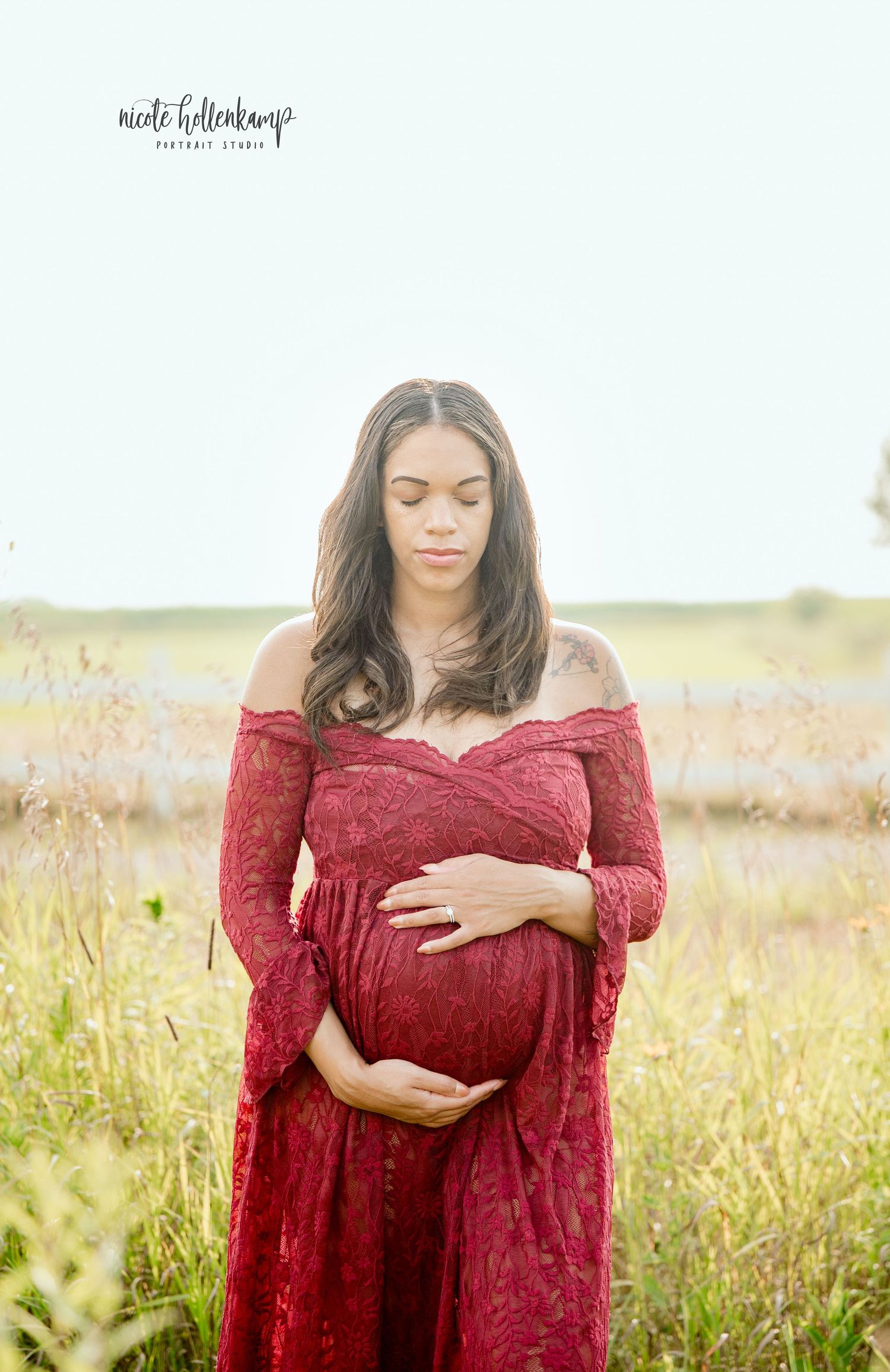 Pregnant woman in red lace off-shoulder gown cradling her baby bump in a sunlit meadow field.