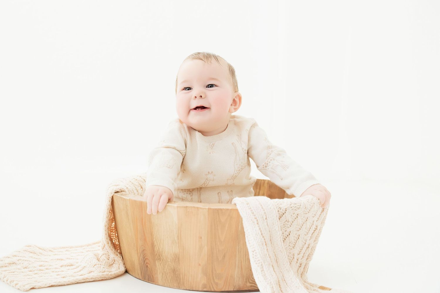 Baby in cream outfit sitting in wooden bowl on white textured blanket.. Minnesota milestone photographer