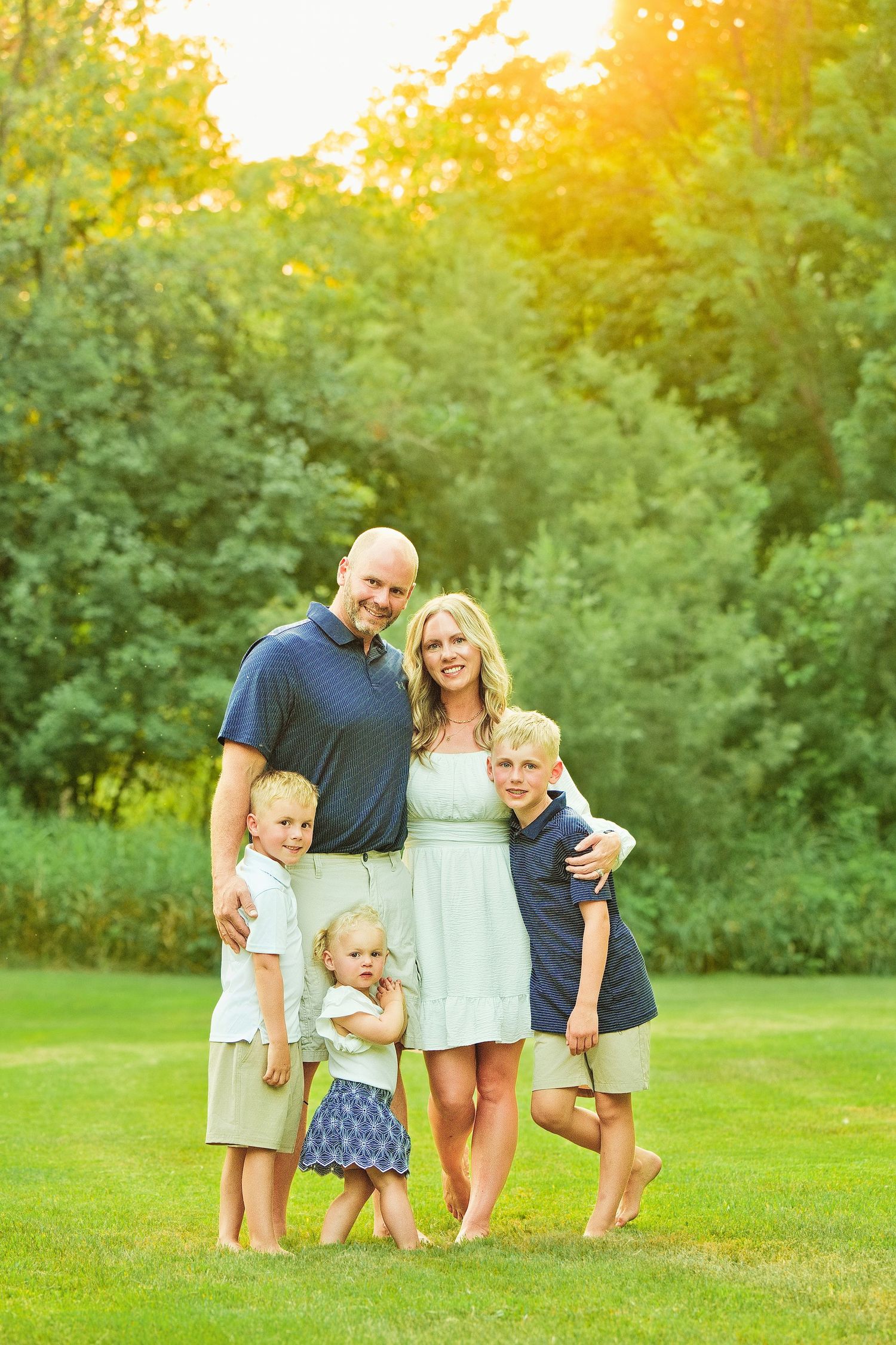 Happy family of five posing outdoors on green grass with sunlit trees in background during golden hour.
